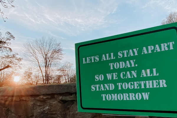Green sign in front of a manmade rock wall that says Let's All Stay Apart Today. So We Can All Stand Together Tomorrow